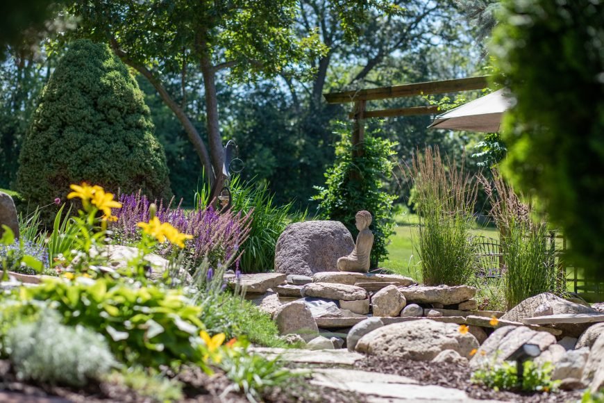 The serenity garden at Denver Psilocybin Sanctuary — stone meditation area with flowering plants, a Buddha statue, and a pergola among lush greenery